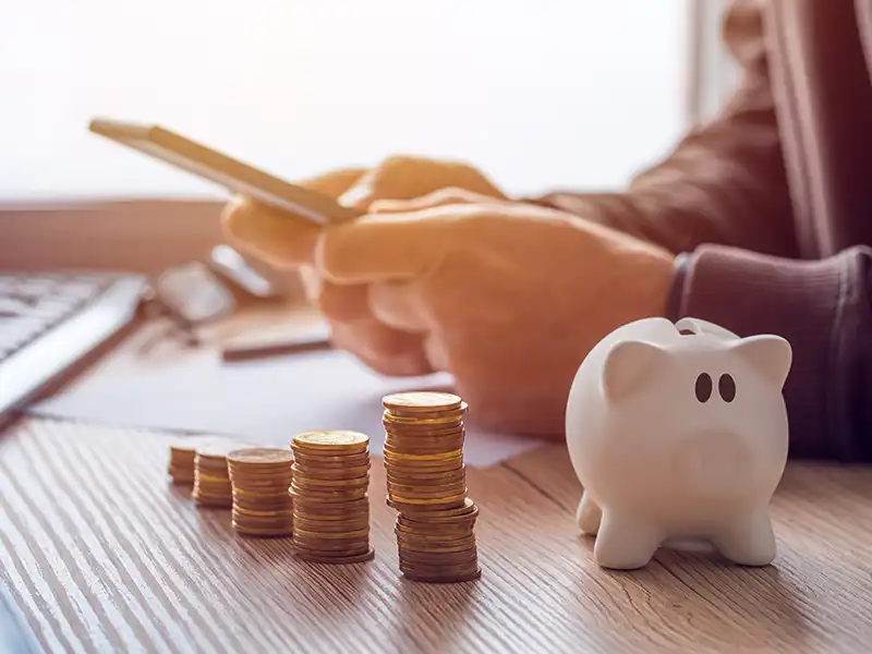 Person using their phone on a desk with a piggybank and coins on top
